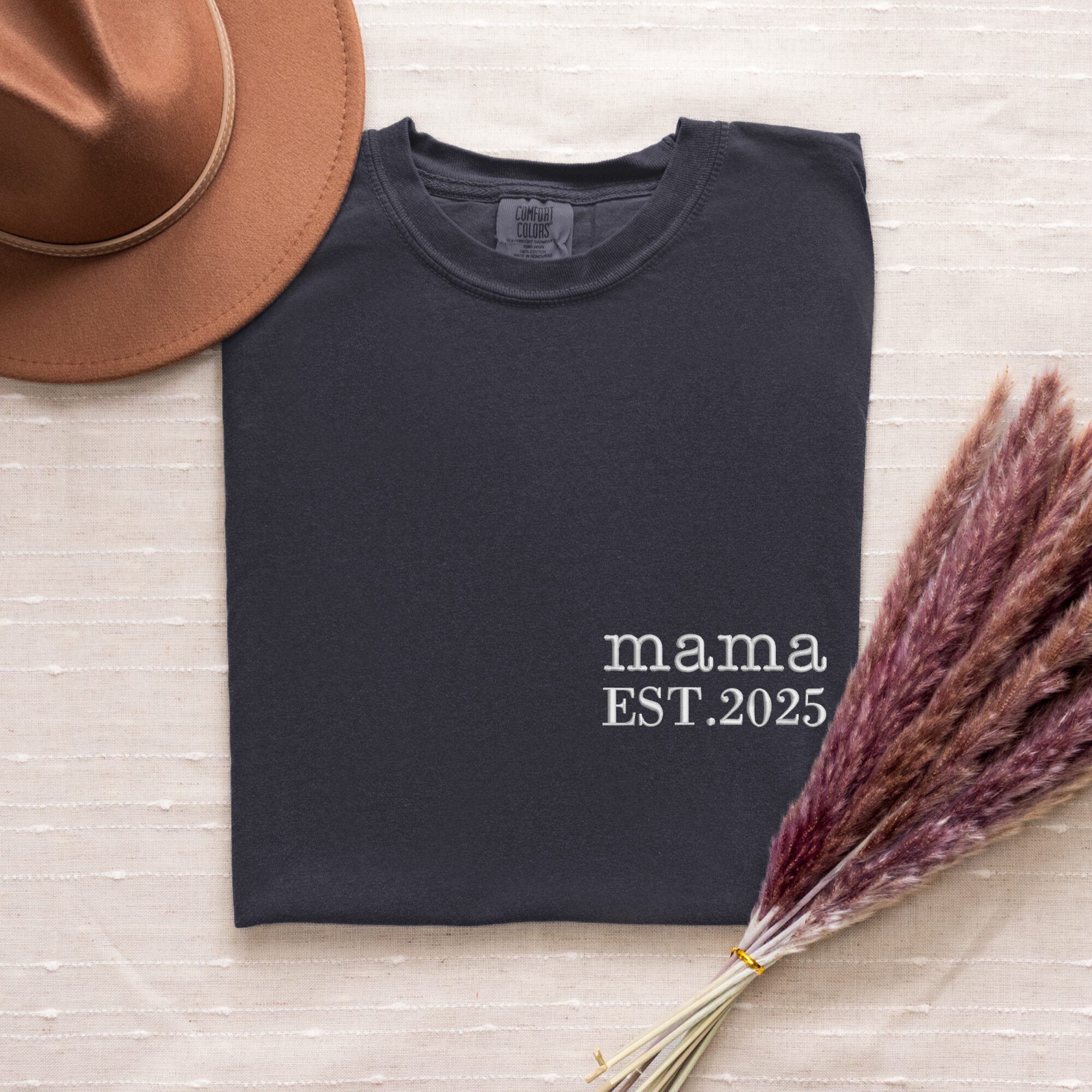 Graphite Comfort Colors Mama T-Shirt folded flat lay with hat and pampas grass, white 'mama EST. 2025'.
